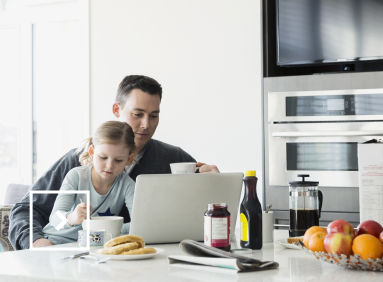 Un homme travaille devant son ordinateur dans sa cuisine, sa fille sur ses genoux.